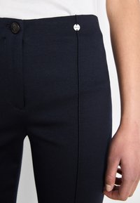 Pantaloni tailleur blu navy con una superficie liscia, dotati di chiusura a bottone e un piccolo dettaglio in argento vicino alla vita.
