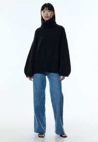 Pull à col roulé noir oversize en tricot avec des manches larges, associé à un jean à jambes larges bleu clair et des sandales noires.
