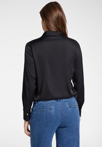 Schwarze Satin-Bluse mit langen Ärmeln und Kragen, kombiniert mit blauen Jeans mit Gesäßtaschen und strukturierter Oberfläche.