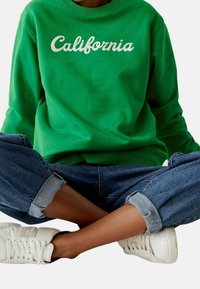 Zelená mikina s bílým vyšitým nápisem "California". Kombinováno s ohrnutými modrými džíny a bílými teniskami, vytvářející ležérní outfit.