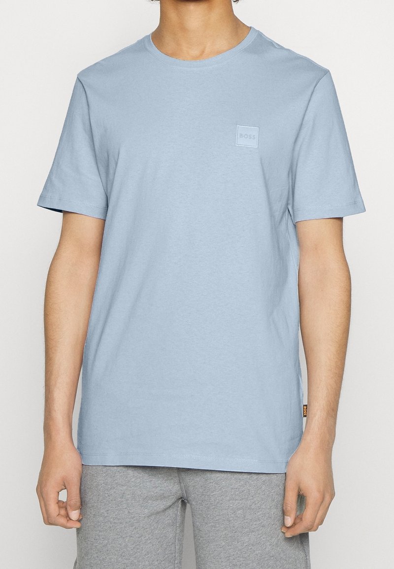 Hellblaues Baumwoll-T-Shirt mit kurzen Ärmeln und rundem Ausschnitt. Auf der Brust befindet sich ein kleines, quadratisches, geprägtes Logo. Glatte Textur.