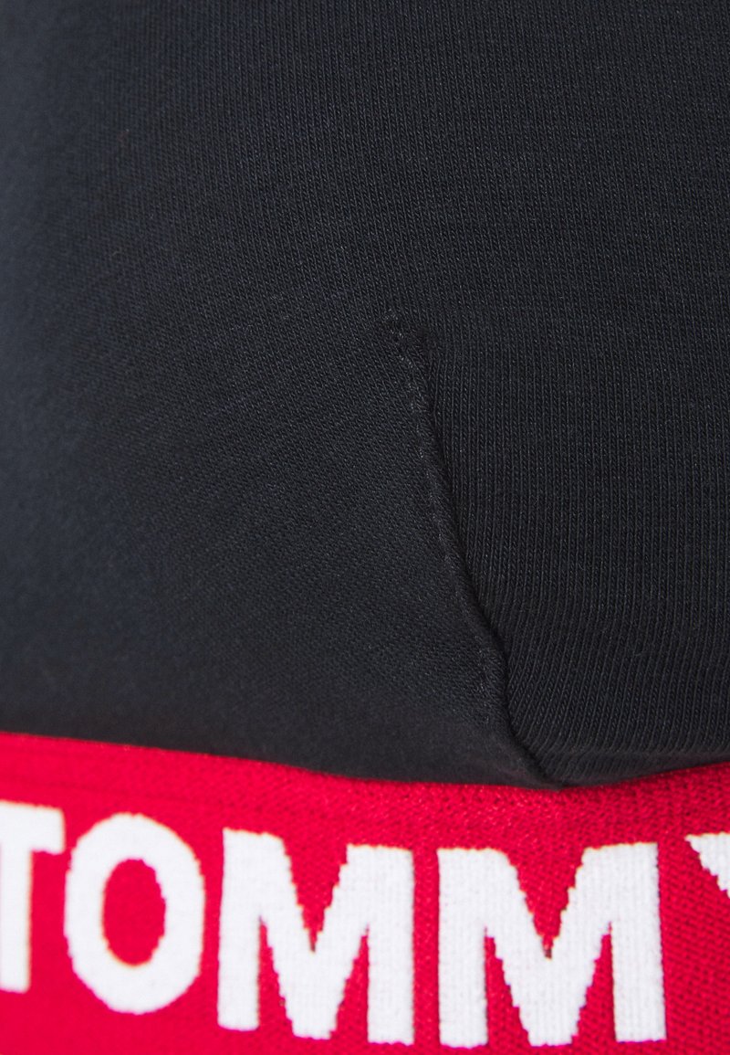 Tejido negro con textura, presentando una banda elástica roja con letras blancas prominentes. Primer plano que muestra detalles del material y del cosido.