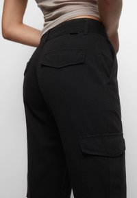 Detailní záběr osoby oblečené v černých cargo kalhotách s kapsami na klopě a zasunutém béžovém triku na jednobarevném pozadí.