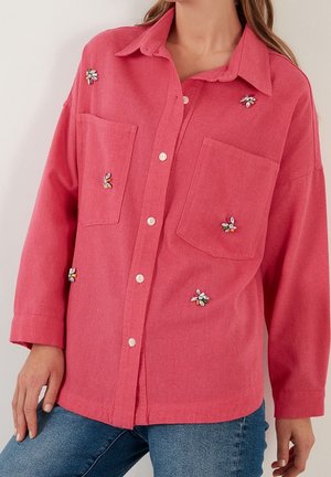 Nainen, jolla on yllään löysä vaaleanpunainen nappipaita, jossa on kukkakoristeiset jalokivisomisteet ja kaksi etutaskua, yhdistettynä sinisiin farkkuihin.