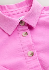 Golier ružovej kordurovej košele so tromi béžovými gombíkmi pozdĺž predného plátu, zobrazujúci textúru rebrovanej látky a detaily švov.
