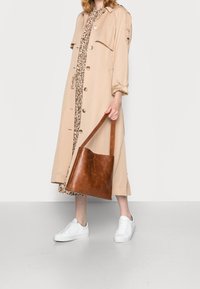Frau in beigem Trenchcoat und gemustertem Kleid, trägt eine braune Lederhandtasche, trägt weiße Sneakers und steht auf einem hellgrauen Boden.