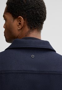 Veste en mélange de laine bleu marine avec un col. Présente un design cousu et un petit logo noir au dos, soulignant la texture.