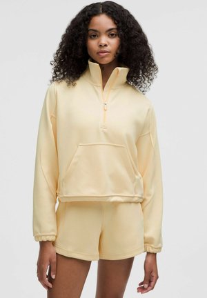 Żółta krótka bluza z wysokim kołnierzem, przednią kieszenią i detalem na zamek; zestawiona z pasującymi szortami, wykonana z miękkiego, elastycznego materiału.