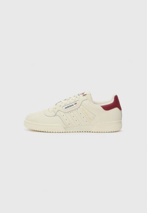 Baskets Adidas crème avec languette rouge au talon, bandes latérales perforées et logo Adidas sur le côté et sur la languette, sur un fond clair uni.