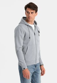 Hoodie zippé gris en mélange de coton, avec une poche avant, des cordons de serrage réglables et un petit patch logo noir sur la poitrine.