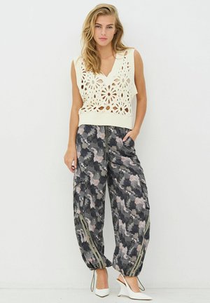 Gehäkeltes, ärmelloses cremefarbenes Oberteil mit floralen Ausschnitten; graue, camouflage-musterte, plissierte Jogginghosen mit seitlichen Reißverschlüssen; kombiniert mit weißen High Heels.