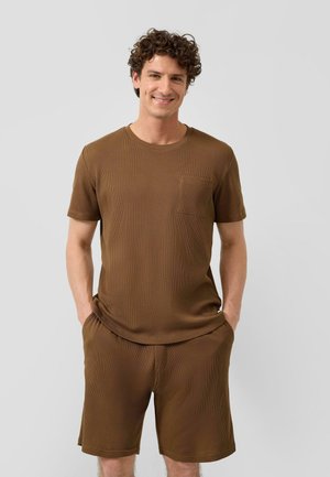 Mann mit lockigem Haar, der lächelt, trägt ein braunes kurzärmeliges Hemd mit Brusttasche und passende braune Shorts, Hände in den Taschen.