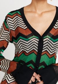 Närbild av en person som bär en knappad V-ringad kofta med ett zigzag-mönster i grönt, brunt, svart och vitt, med enfärgade svarta manschetter.