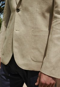Svijetlosmeđi laneni blazer s dvostrukim kopčanjem, izrezom na reverima, zakrpama te suptilnom teksturom, uparen s tamnim hlačama s uzorkom.