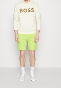 Sudadera crema con la palabra "BOSS" en letras marrones, combinada con pantalones cortos verdes brillantes. Zapatillas altas blancas y calcetines blancos completan el look.