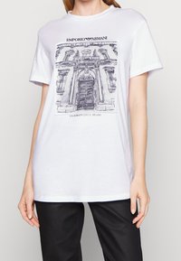 Bílý bavlněný tričko s šedou ilustrací fasády budovy a textem "EMPORIO ARMANI" a "VIA BORGONUOVO 11, MILANO."