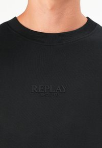 Black knit sweatshirt featuring a ribbed crew neck and a woven "REPLAY SINCE 1981" logo embossed on the front. Soft texture.