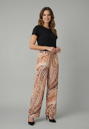 Femme souriante debout, les mains jointes, portant un haut noir à manches courtes, un pantalon large beige à motifs et des chaussures à talons noirs.
