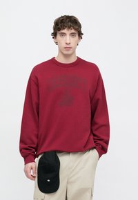 Κόκκινο φούτερ με στρογγυλή λαιμόκοψη και υφή με ανάγλυφο φινίρισμα, με σκούρο λογότυπο τυπωμένο. Συνδυασμένο με μπεζ παντελόνι cargo και μαύρο καπέλο που ακουμπά στη μέση.