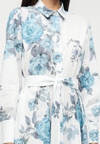 Φλοράλ φόρεμα πουκάμισο σε λευκό με μπλε φλοράλ σχέδιο, με γιακά με κουμπιά, μακριά μανίκια και ζώνη στη μέση που δένει.