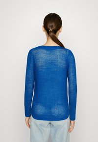 Kvinna med brunt hästsvans som bär en genomskinlig blå stickad tröja och ljusblå jeans, stående vänd mot en vit bakgrund.