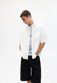 Chemise blanche à manches courtes avec des boutons et une poche poitrine, portée par-dessus une chemise grise et assortie à un short en denim noir. Texture lisse.