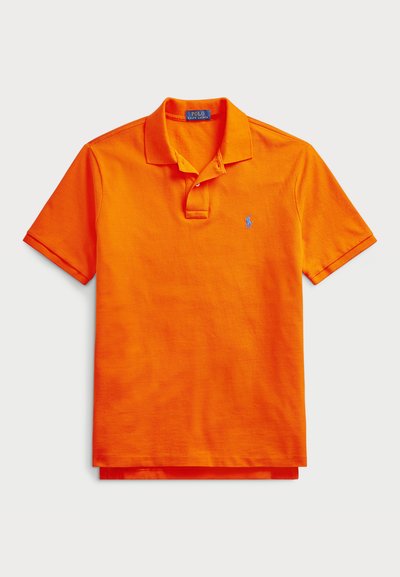 Oransje poloskjorte laget av bomull; har krage, to-knapps placket, og liten blå brodert logo på venstre bryst. Korte ermer.