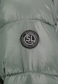Groene pufferjas met een glanzende, gladde textuur. Bevat een ronde zwarte logo patch met het label "SL STUDIO" en details op de stof.