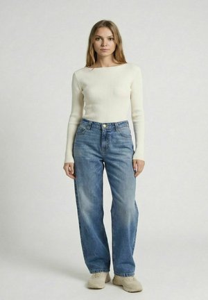 Žena stojaca, oblečená v bielom rebrovanom tričku s dlhým rukávom, voľné modré džínsy a béžové tenisky na jednoduchom svetlom pozadí.