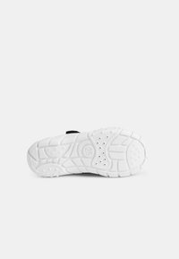 Semelle de chaussure de sport blanche avec un motif texturé et des découpes pour une meilleure adhérence, dotée d'une sangle noire sur la partie supérieure pour un maintien sécurisé.
