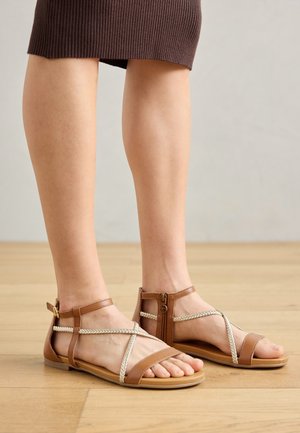 Woman wearing brown flat sandals with braided straps, standing on a wooden floor, wearing a ribbed brown skirt.