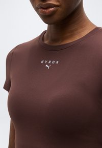 Γυναίκα που φοράει στενή καφέ αθλητική μπλούζα με λευκό λογότυπο "HYROX" και λογότυπο Puma στο στήθος, μπροστά σε απλό φόντο.