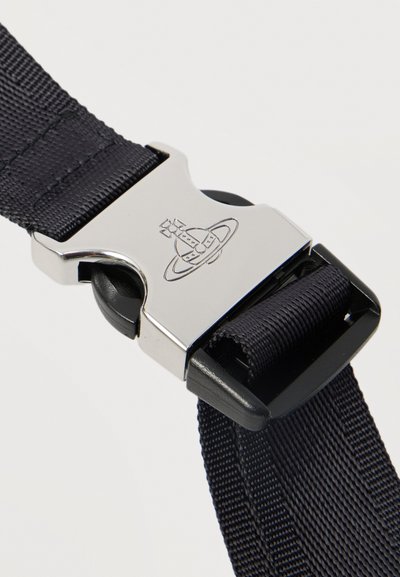 Μεταλλική πόρπη με κυκλικό έμβλημα που συγκρατεί δύο μαύρα υφασμάτινα πλεκτά λουριά πάνω σε ανοιχτό φόντο.