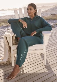 Türkiser Kapuzen-Overall mit Reißverschluss, mit dem Aufdruck "ELB SAND" auf der Hose. Modell sitzt auf einem weißen Holzstuhl auf einer Terrasse.