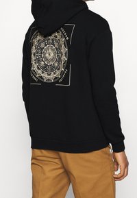 Sweat à capuche noir avec un grand motif doré complexe au dos, comprenant des designs abstraits et du texte. Matière en mélange de coton.