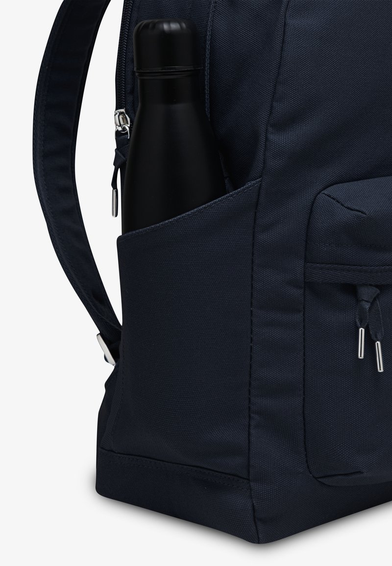 Mochila azul marino hecha de tejido texturizado, con un bolsillo lateral que sostiene una botella de acero inoxidable negra y herrajes en plata con contraste.