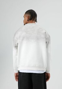 Witte trui met een grijze verloopprint, ronde hals en geribbelde boorden; gedragen over een zwarte broek. Achterkant heeft een vervaagde textuur.