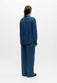 Femme aux longs cheveux bruns portant une chemise en denim ample et un pantalon large en denim, debout de dos sur un fond blanc uni.