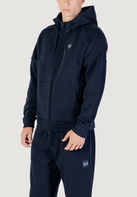 Navy Zip-Hoodie aus weichem Stoff, mit einer geräumigen Kapuze, seitlichen Taschen und einem kleinen Logo-Patch auf der Brust.