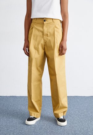 Wide-leg, high-waisted yellow cotton pants with pleats, featuring a front button and side pockets, paired with black and white sneakers.