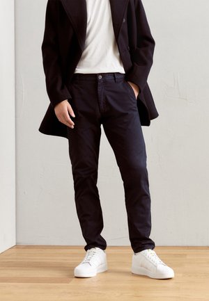 Homme portant un pantalon noir, des baskets blanches, une chemise blanche et un blazer noir, debout sur un sol en bois face à un mur clair.