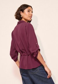 Veini värvi, kerge pluus, millel on kogutud talje ja kolmeveerandi pikkusega varrukad, klassikalise krae ja peente nööbidenähtavustega.