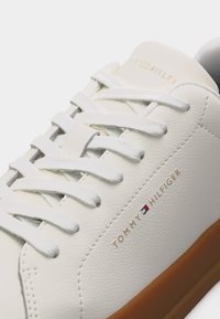 Λευκό δερμάτινο αθλητικό παπούτσι με λευκά κορδόνια, καφέ σόλα και λογότυπο "Tommy Hilfiger" με λεπτομέρεια σημαίας στο πλάι.