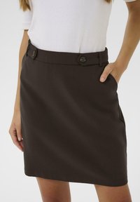 Jupe brune avec une coupe droite, deux poches avant et une taille à boutons. Le matériau a une texture lisse avec un subtil éclat.