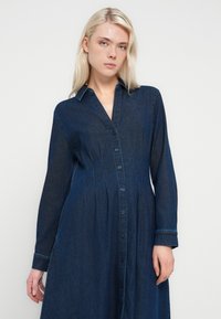 Denimskjortekjole i mørkeblå. Har knapper foran, lange ærmer, krave og en struktureret silhuet med sidelommer.