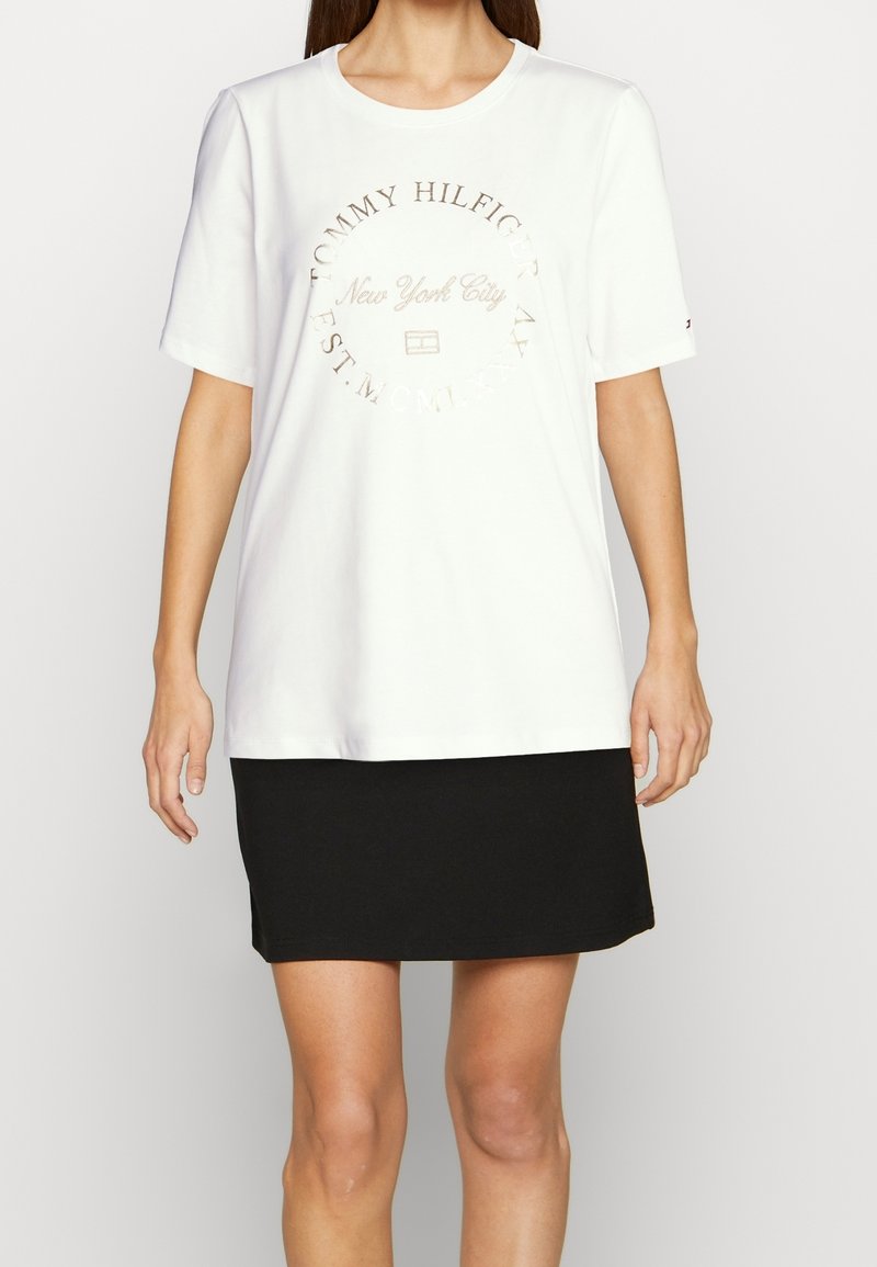 Camiseta blanca de algodón con un logo circular en dorado metálico que dice "New York City". Combinada con una falda negra ajustada.