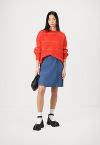 Red knitted sweater with striped pattern, paired with a blue denim skirt. Model wears black chunky shoes and holds a black bag.