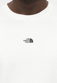 Στενή λήψη ενός ατόμου που φοράει λευκό πουκάμισο με μαύρο λογότυπο The North Face στο στήθος και στρογγυλή λαιμόκοψη.