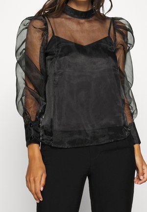 Zwarte doorzichtige organza blouse met opgeblazen lange mouwen, knoopmanchetten en een aansluitend zwart hemd eronder, gecombineerd met zwarte broek.