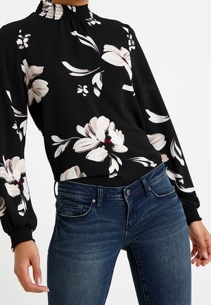 Femme portant un chemisier noir à manches longues avec des imprimés floraux blancs et un jean en denim bleu sur un fond uni.
